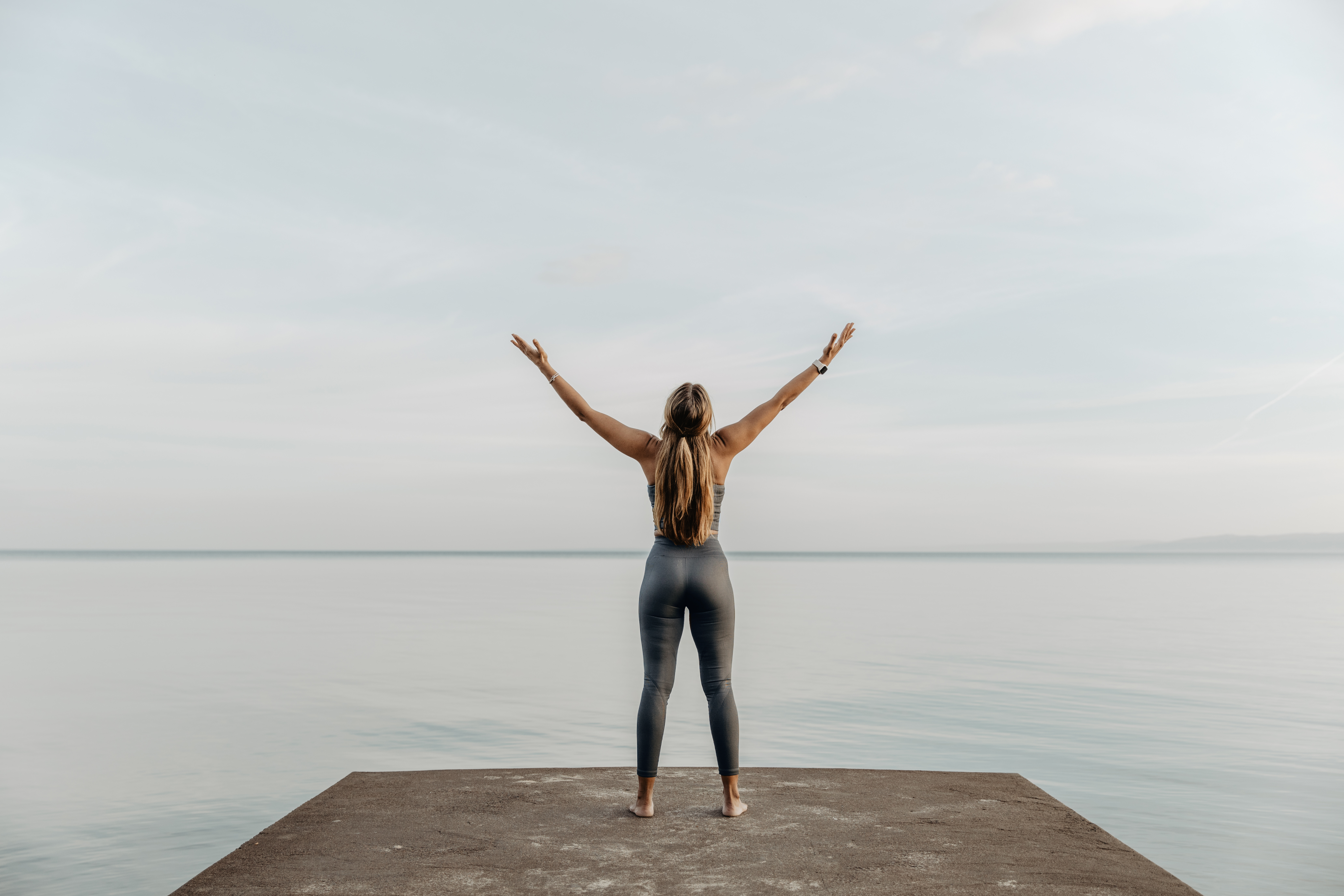 Vatten och himmel. En kvinna står ute på bryggan och sträcker upp armarna mot himlen. Bilden går i lugna, grå toner.