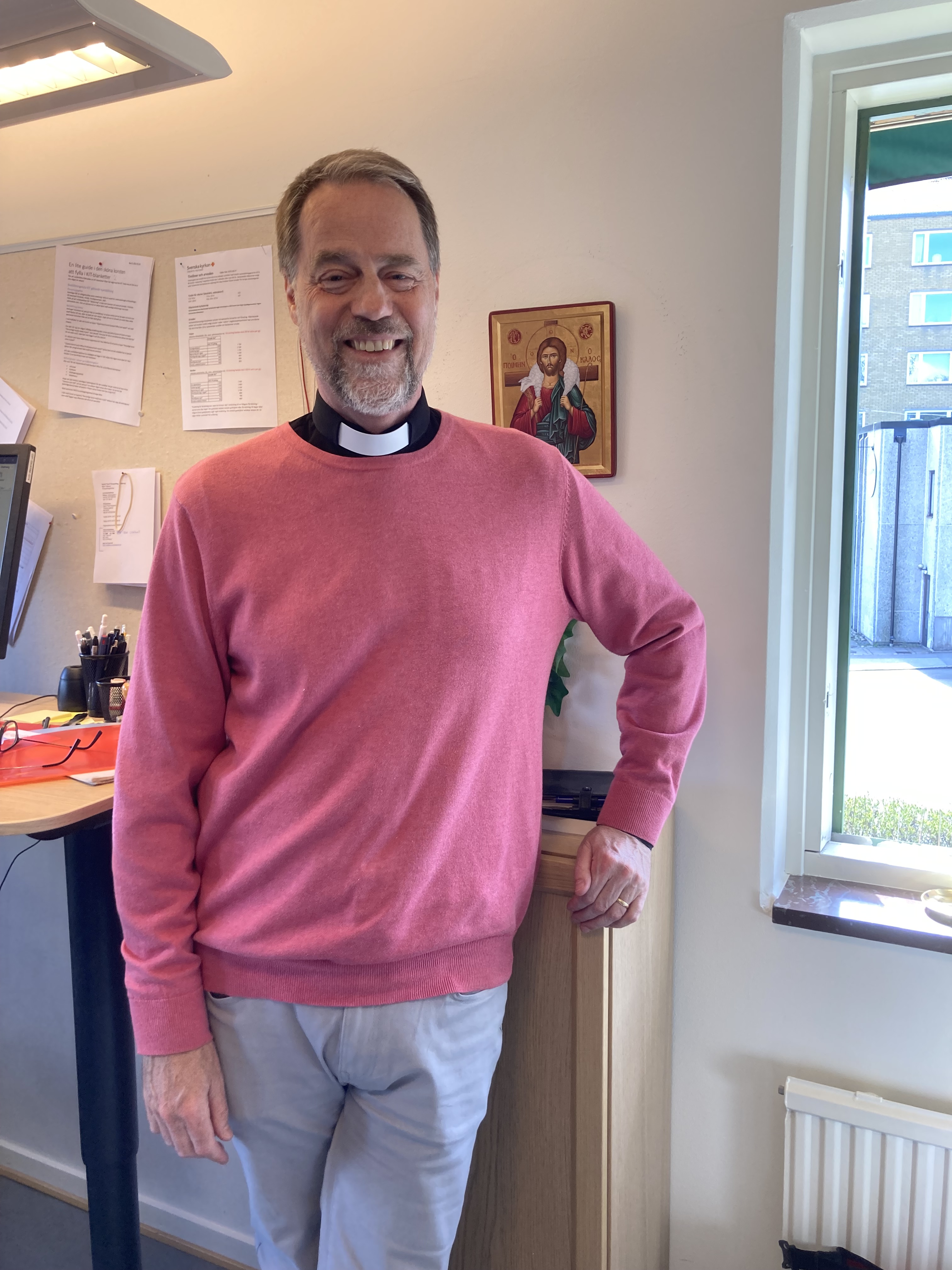 Björn Nilsson, stående i en kontorsmiljö, klädd i prästskjorta och rosa tröja.