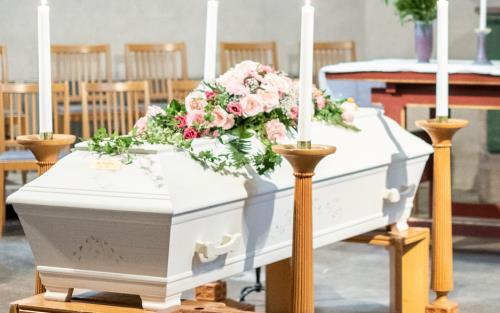 En kyrksal med en vit kista med rosa rosor på locket står till vänster i bild. På andra sidan om kistan står en präst i liturgiska kläder och med nerböjt huvud. I förgrunden syns delar av kyrkbänkar i trä.