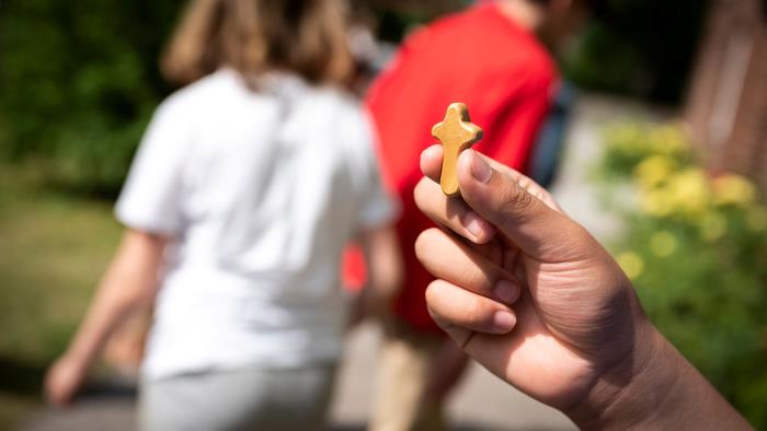 En hand som håller i ett litet träkors. I bakgrunden två personer som går iväg.