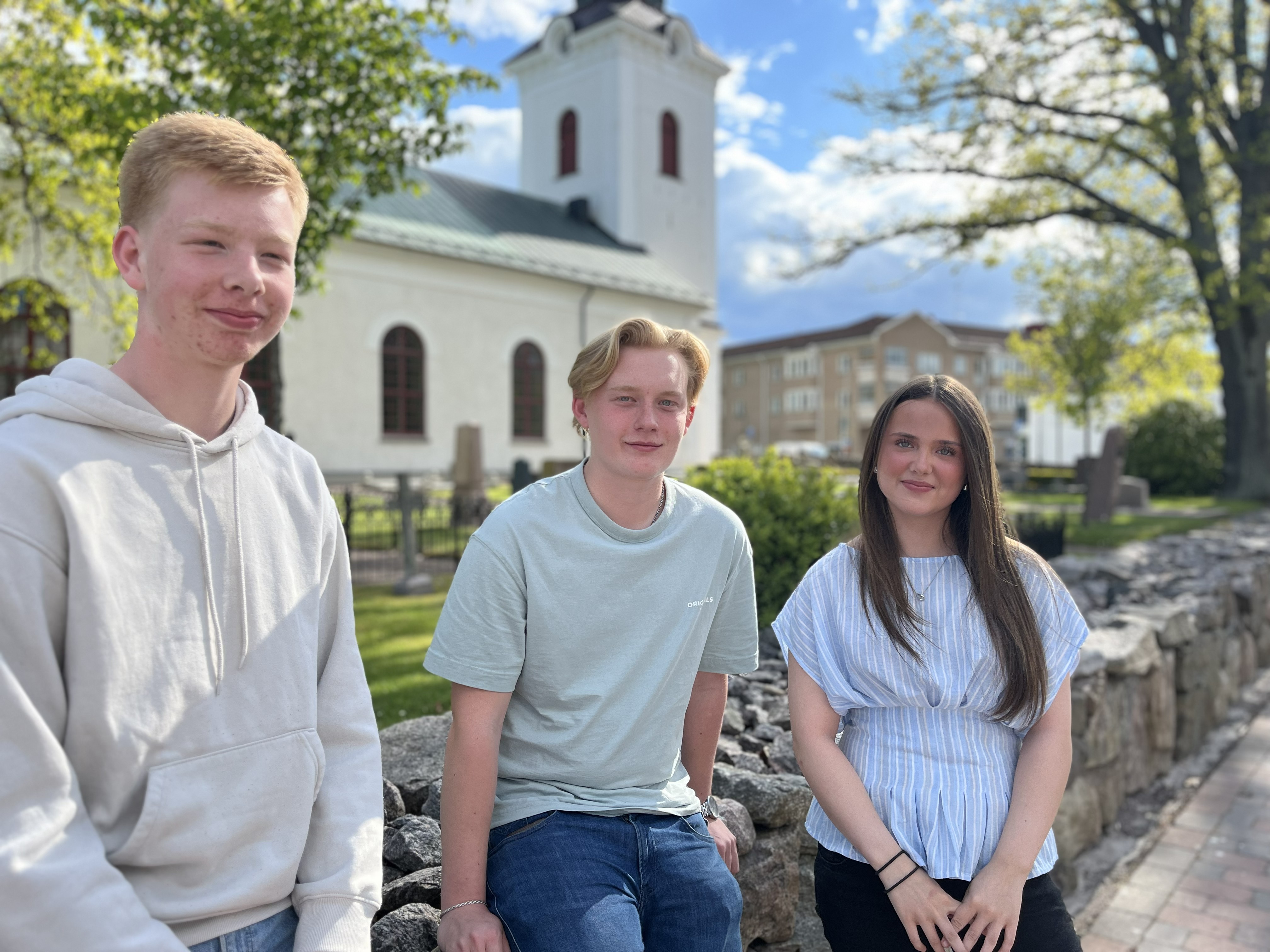 Tre ungdomar bredvid varandra, bakom dem syns en kyrka.