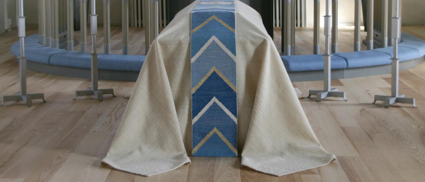 En kista står framför altaret i Tidaholms kyrka, med ett vitt bårtäcke och en blå, vit och silvrig bård ovanpå.