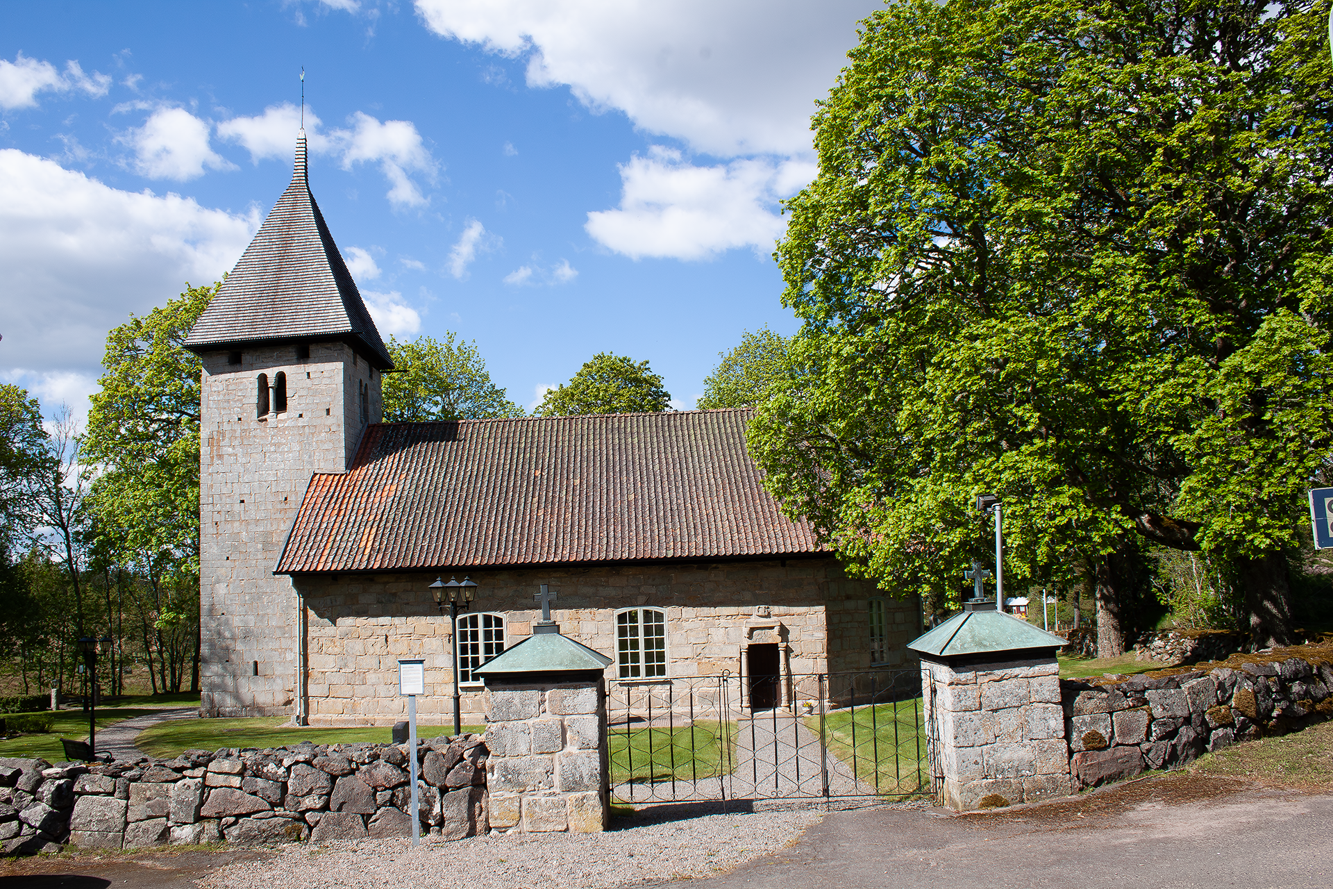 En stenkyrka med torn och långhus, runt omkring är lummiga träd och framför kyrkan en stenmur med tillhörande grind.