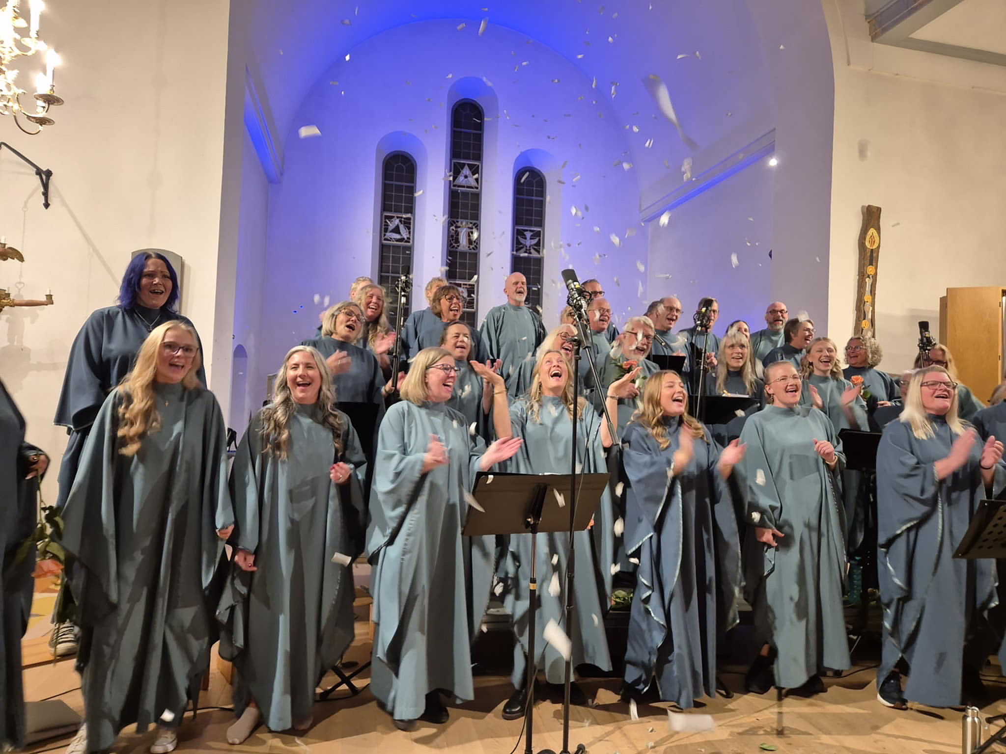 Körmedlemmar klädda i turkosa körkåpor, sjunger med glädje i en kyrka, med konfetti i luften.