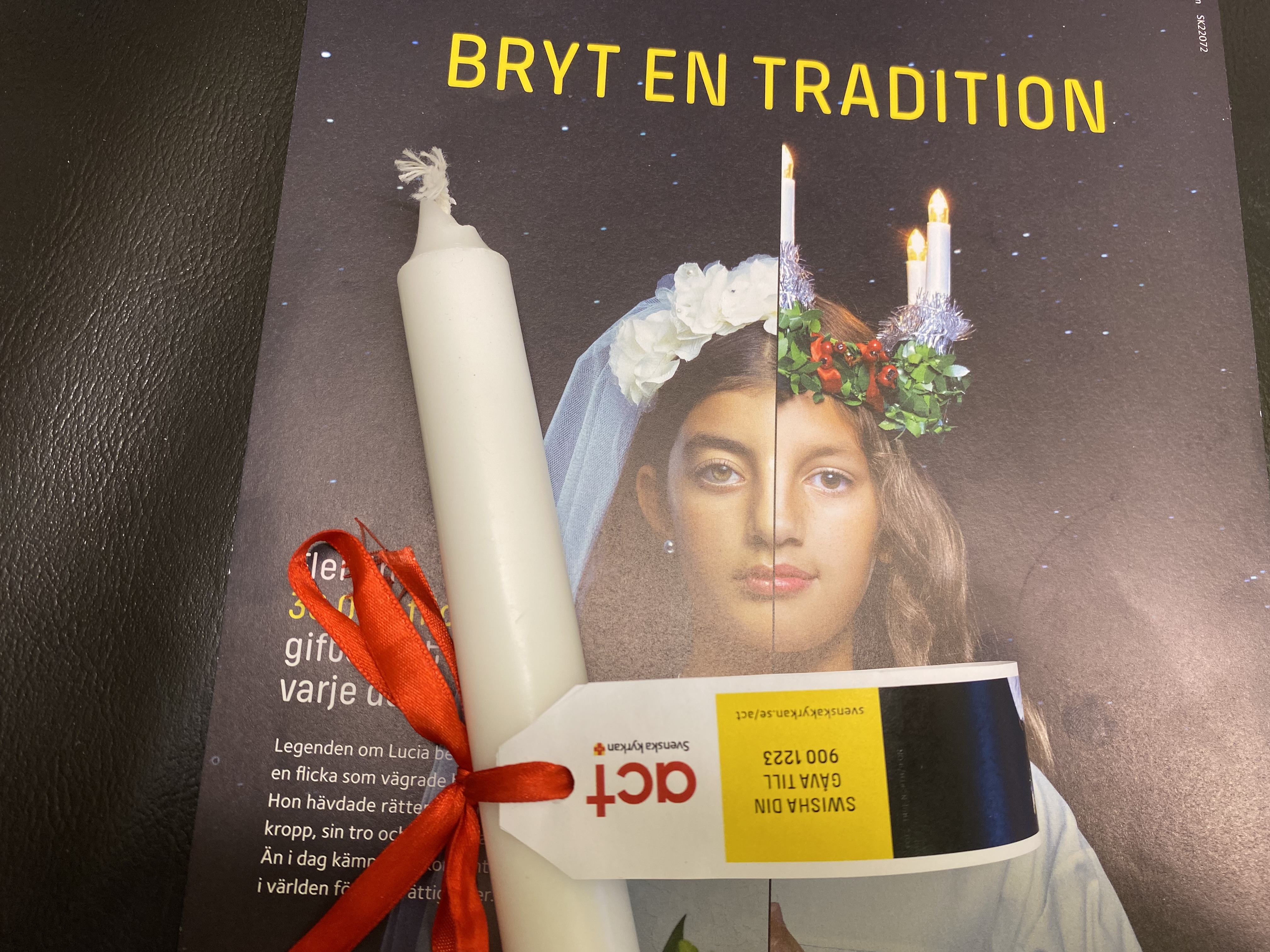 Ett vitt ljus med rött band och en etikett med information om Act Svenska kyrkans julinsamling samt en affisch från Act Svenska kyrkan med texten "Bryt en tradition" och kampanjbilden för julinsamlingen 2022.