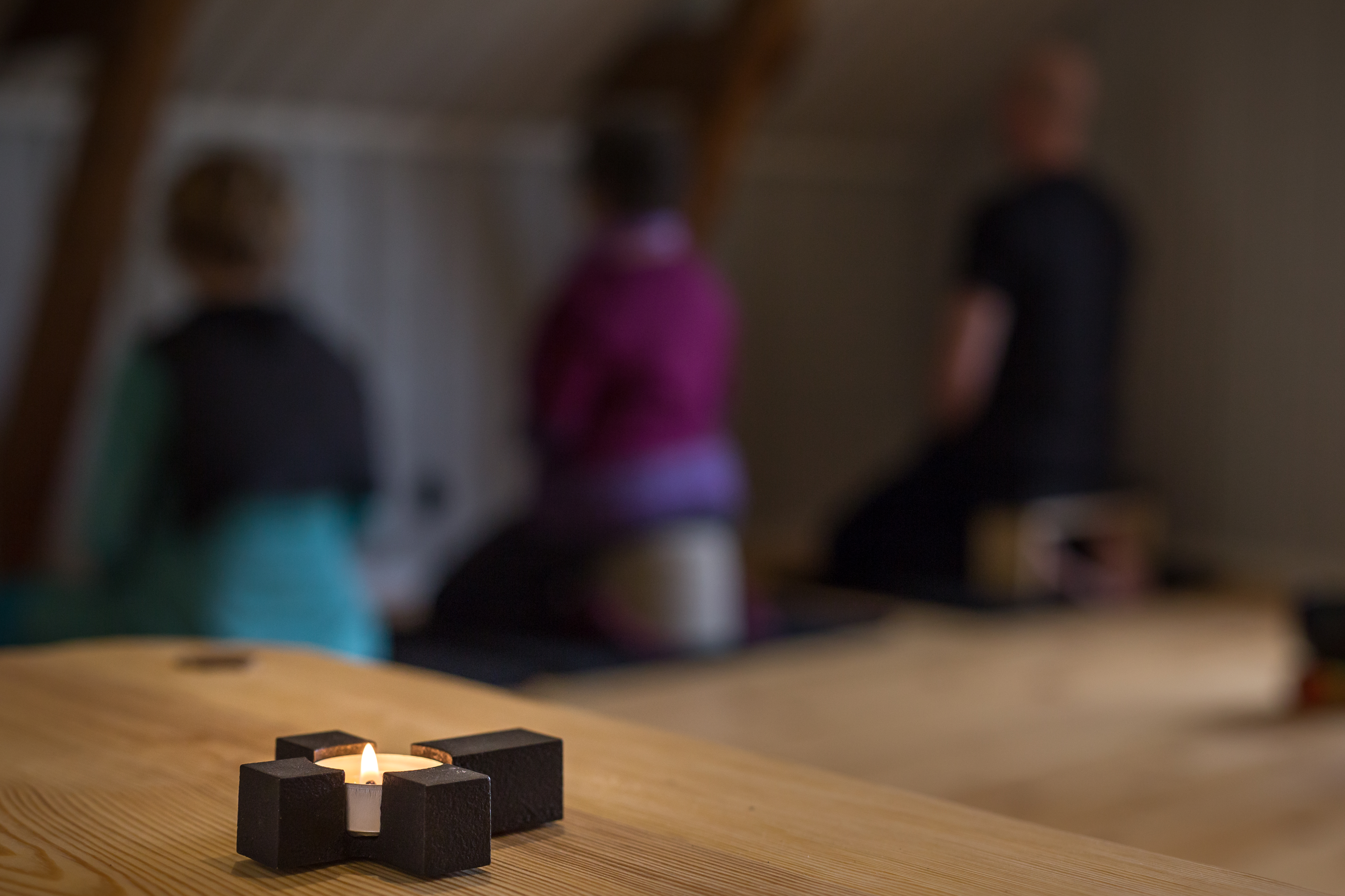 Ett kors med ett värmeljus i förgrunden och i bakgrunden sitter tre personer med ansiktet på väggen och mediterar.