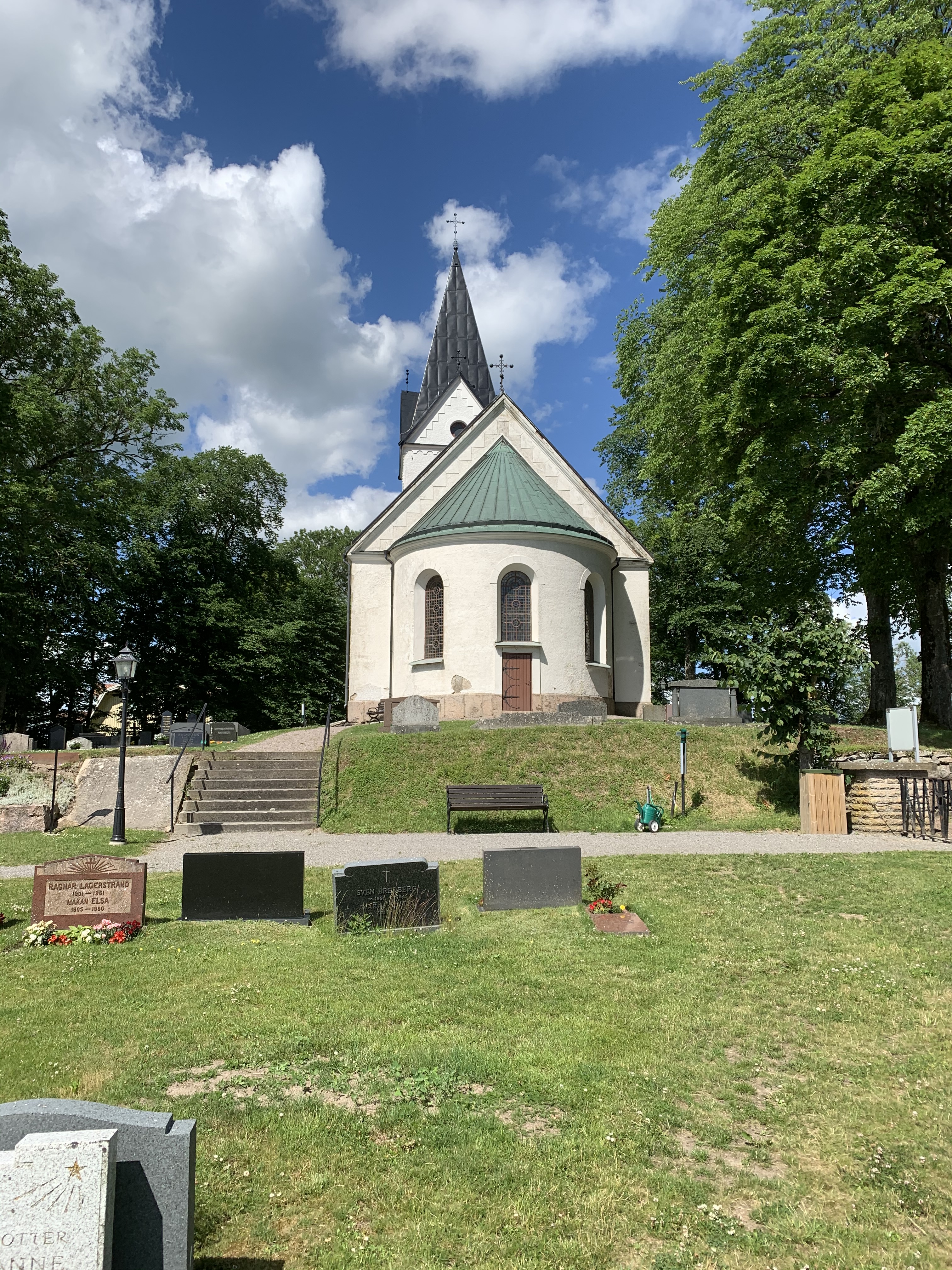 Kymbo kyrka sett bakifrån, runt omkring syns kyrkogården, grönt gräs och gröna, lummiga träd samt blå himmel.