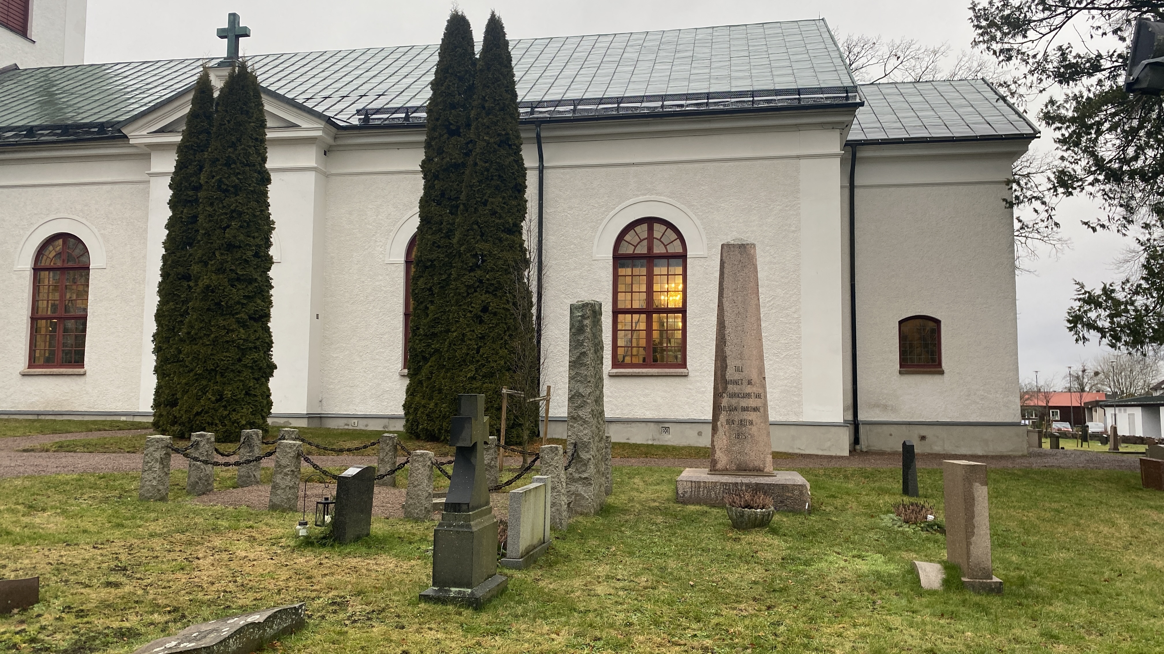 En del av en kyrka och framför står en rest minnessten på kyrkogården.