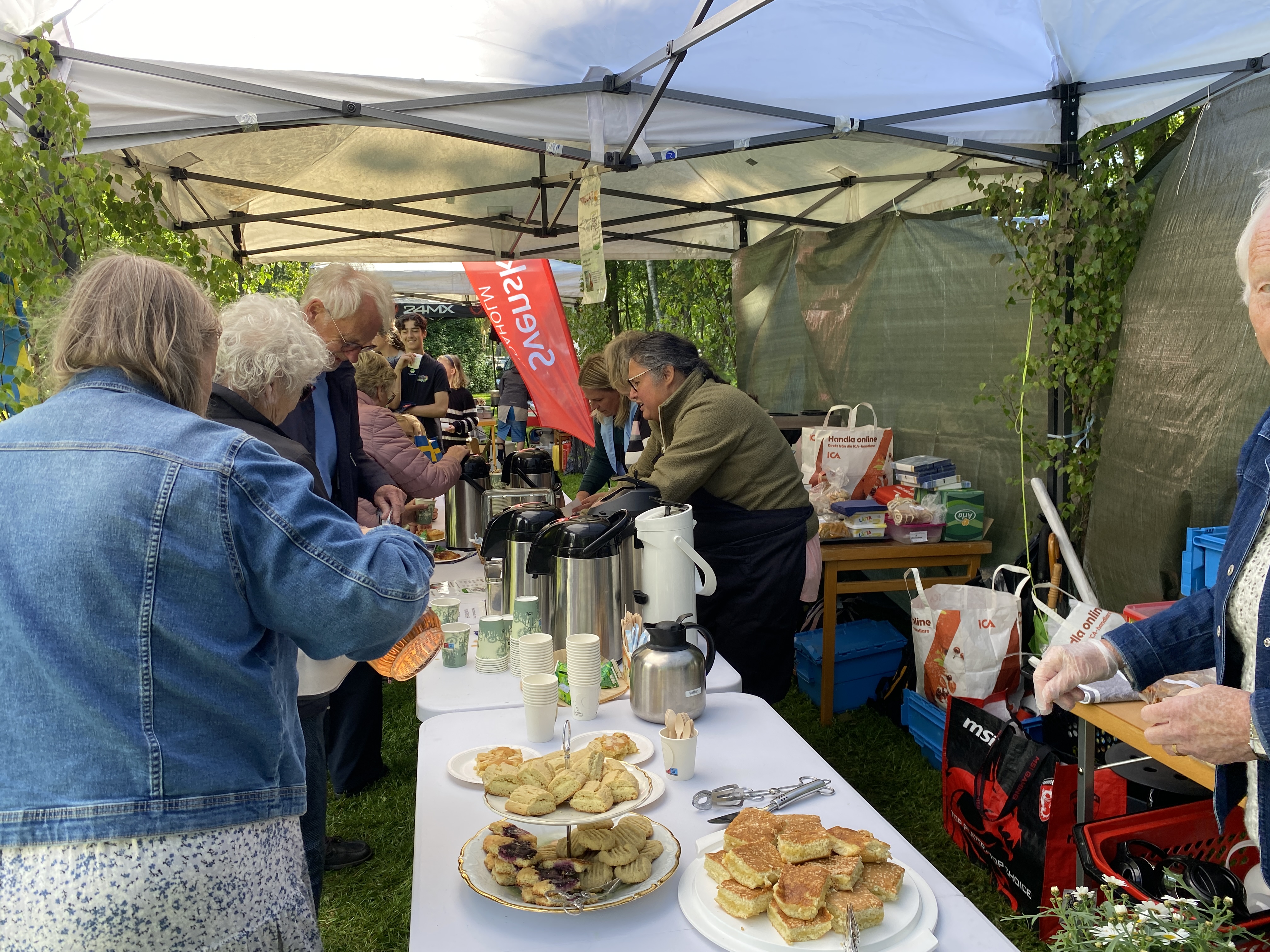 Några personer blir serverade kaffe under ett tält, på ett bort finns även kakor.