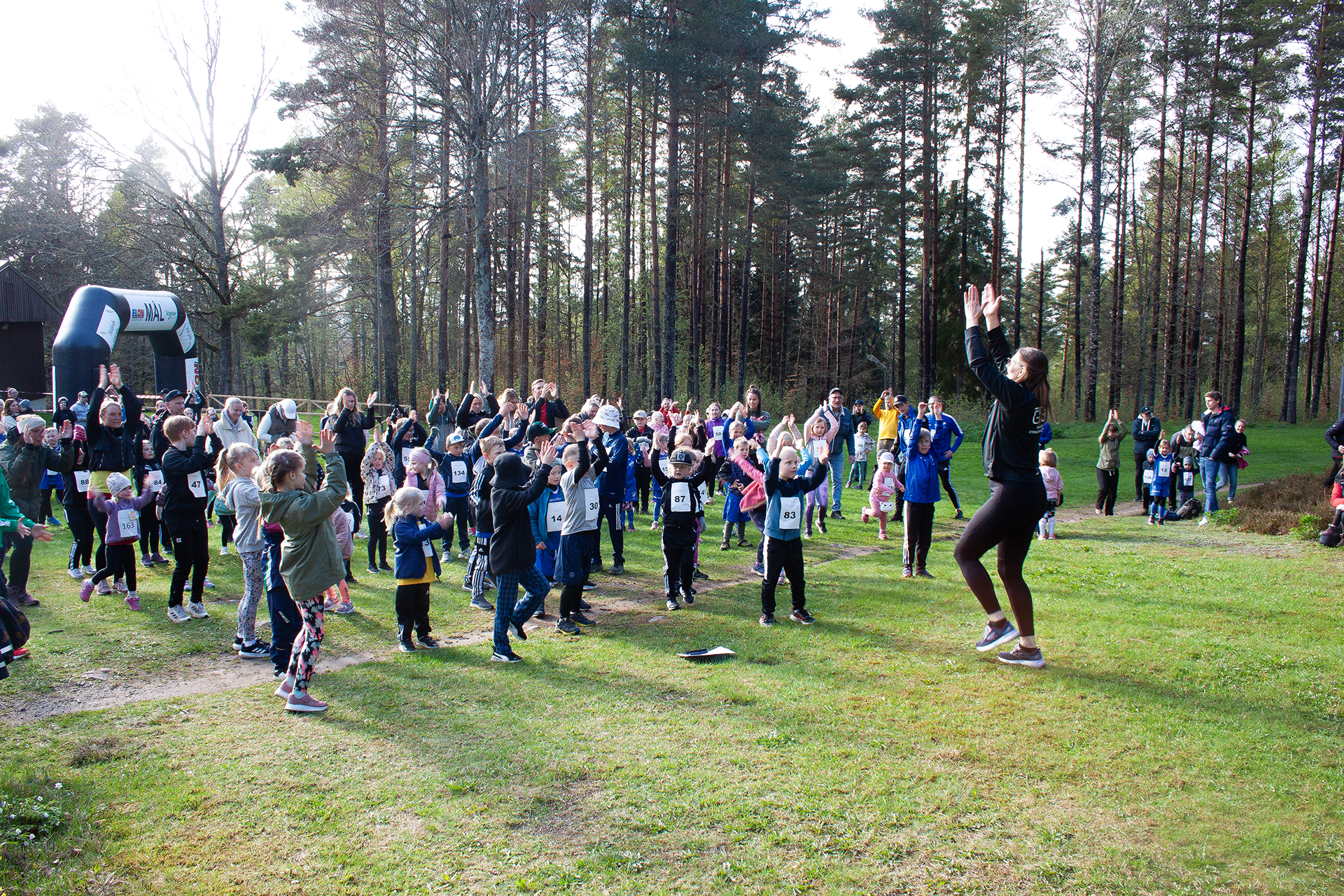 Ett gäng barn, ungdomar och vuxna håller händerna i luften, de tittar på en kvinna i träningskläder.