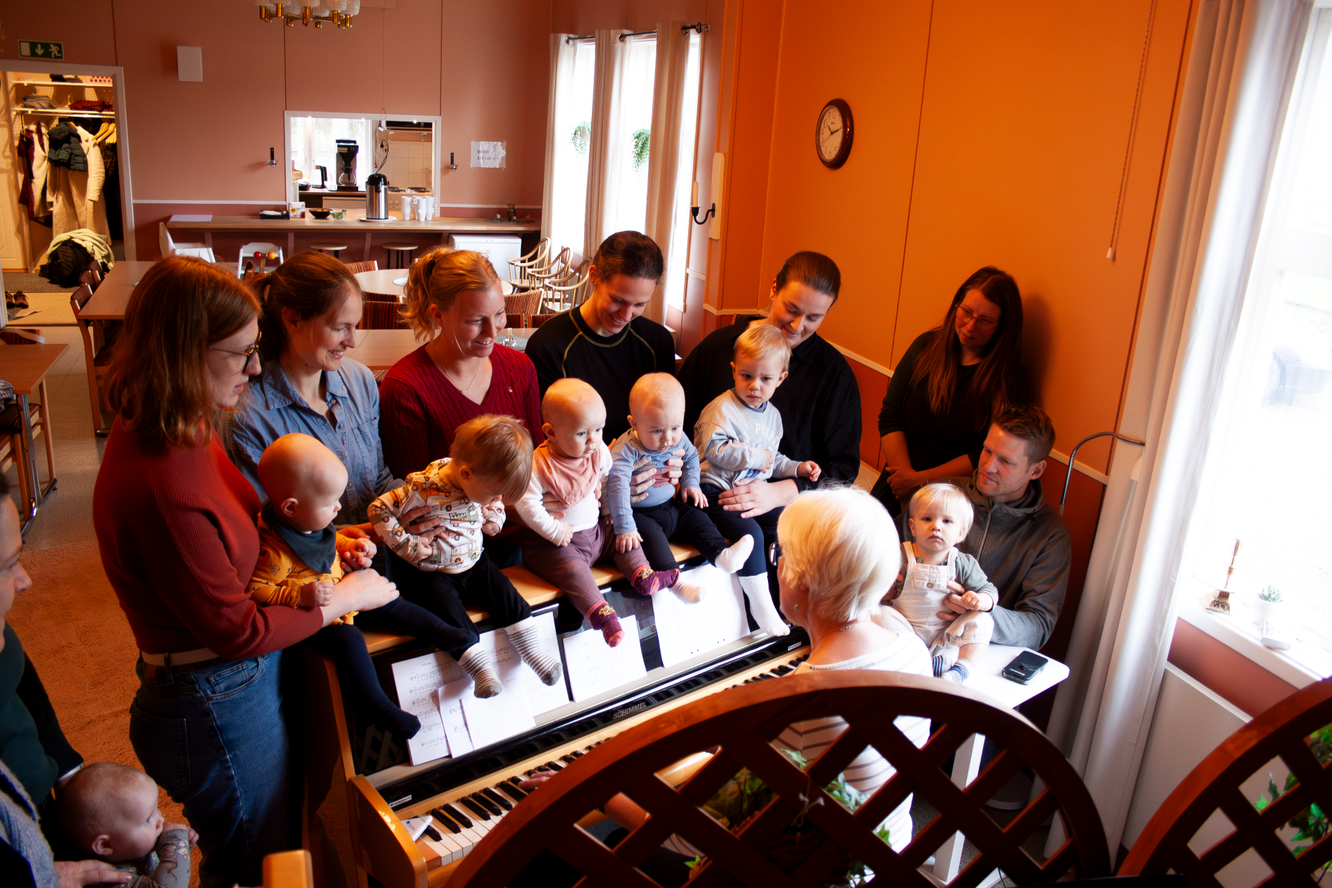 Ett gäng små barn sitter bredvid varandra på ett piano, bakom dem står deras föräldrar som håller i sina barn. Framför dem sitter en kvinna och spelar på pianot.