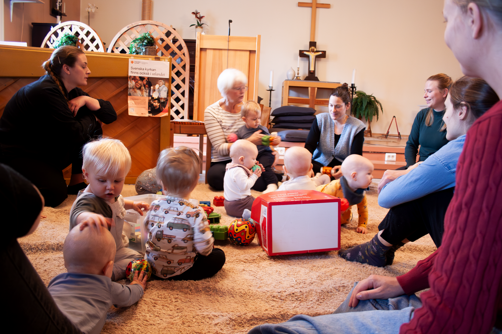 Några barn och vuxna sitter på golvet i en församlingssal och leker och pratar.