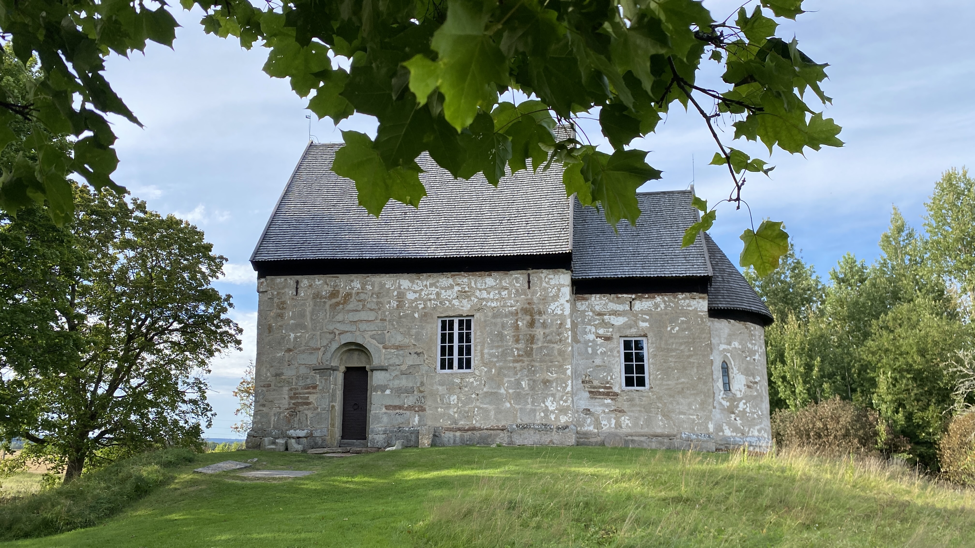 En stenkyrka, med gröna löv ovanför i förgrunden och grönskande träd och buskar i bakgrunden.