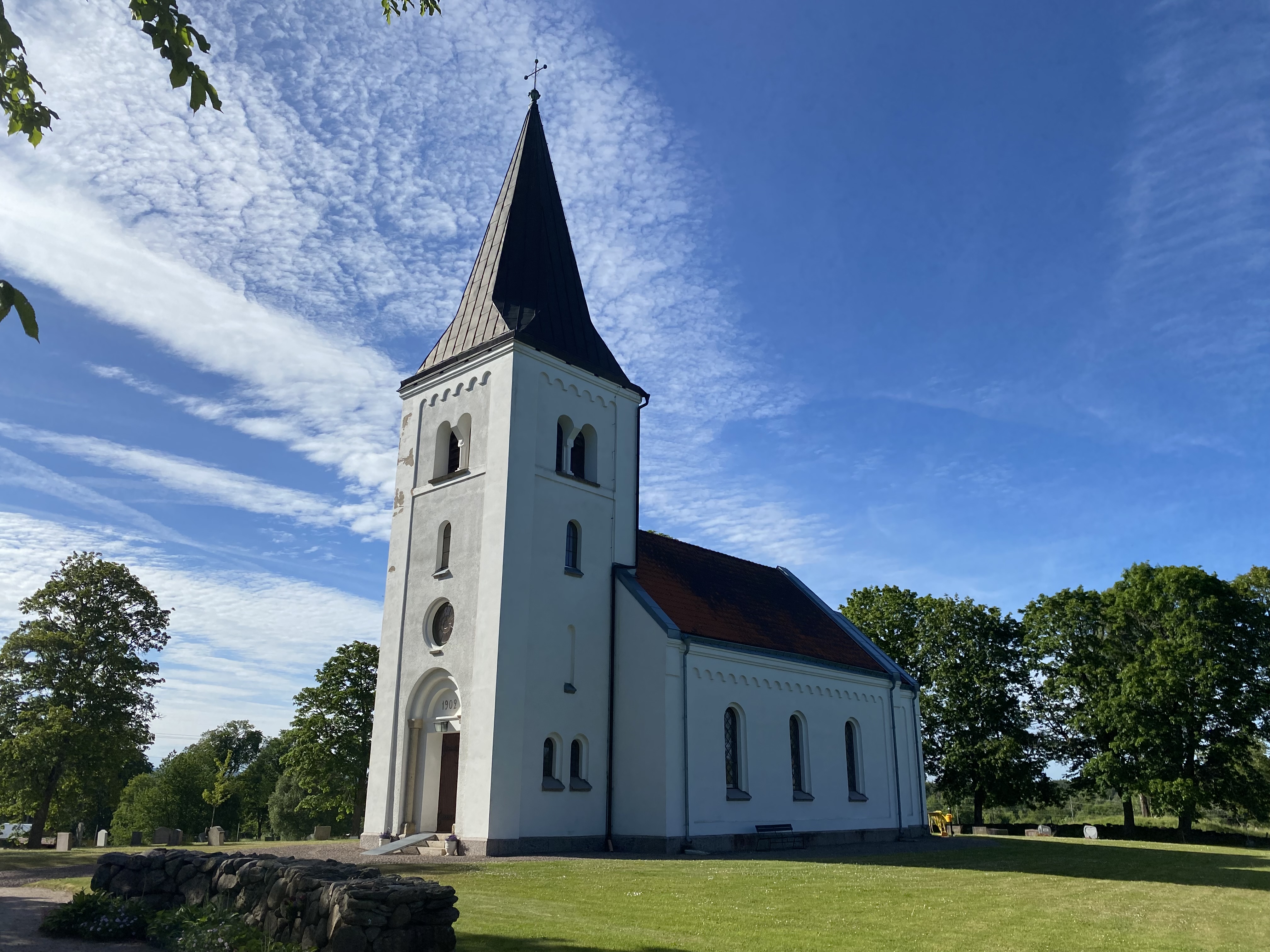 Suntaks kyrka i skugga med grönskande träd runt omkring, blå himmel och grönt gräs.