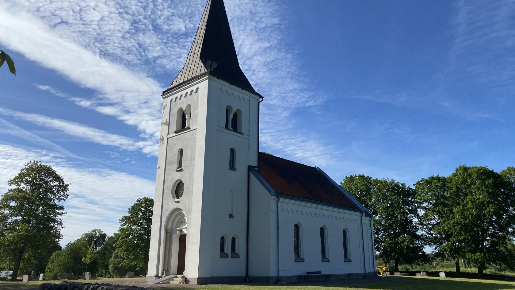 Suntaks kyrka i skugga med grönskande träd runt omkring, blå himmel och grönt gräs.