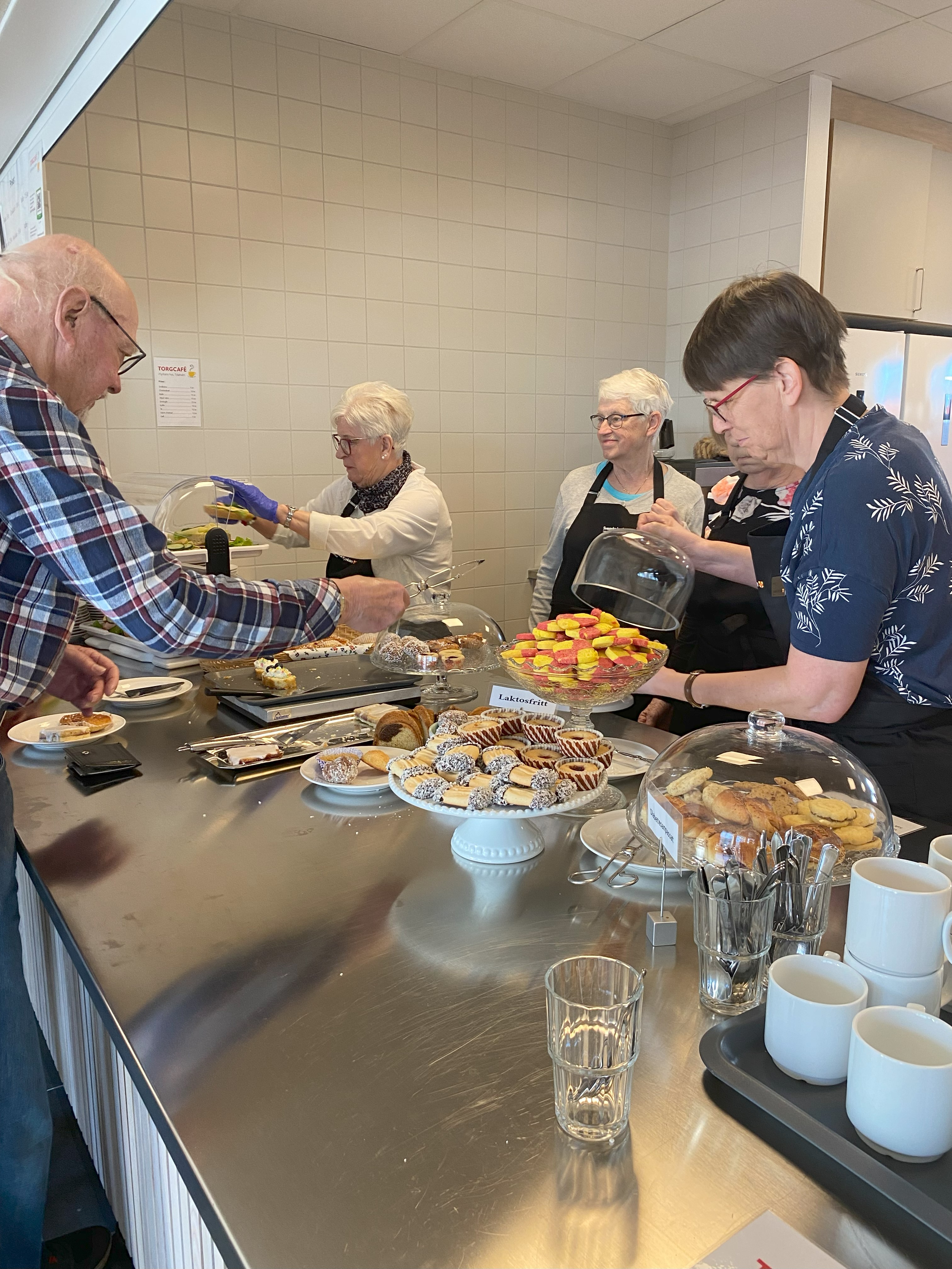 Volontärer i svarta förkläden med Svenska kyrkan Tidaholms logga på hjälper torgcafékunder.