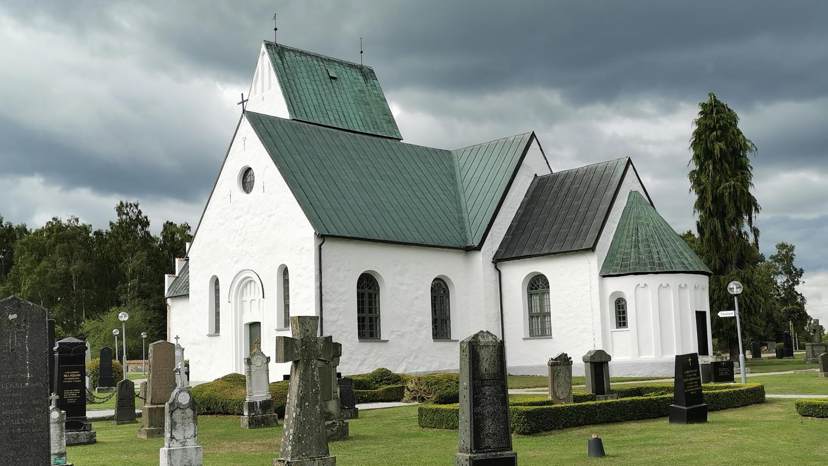 Köpinge kyrka med gröna träd i bakgrunden och svarta moln på himmelen.