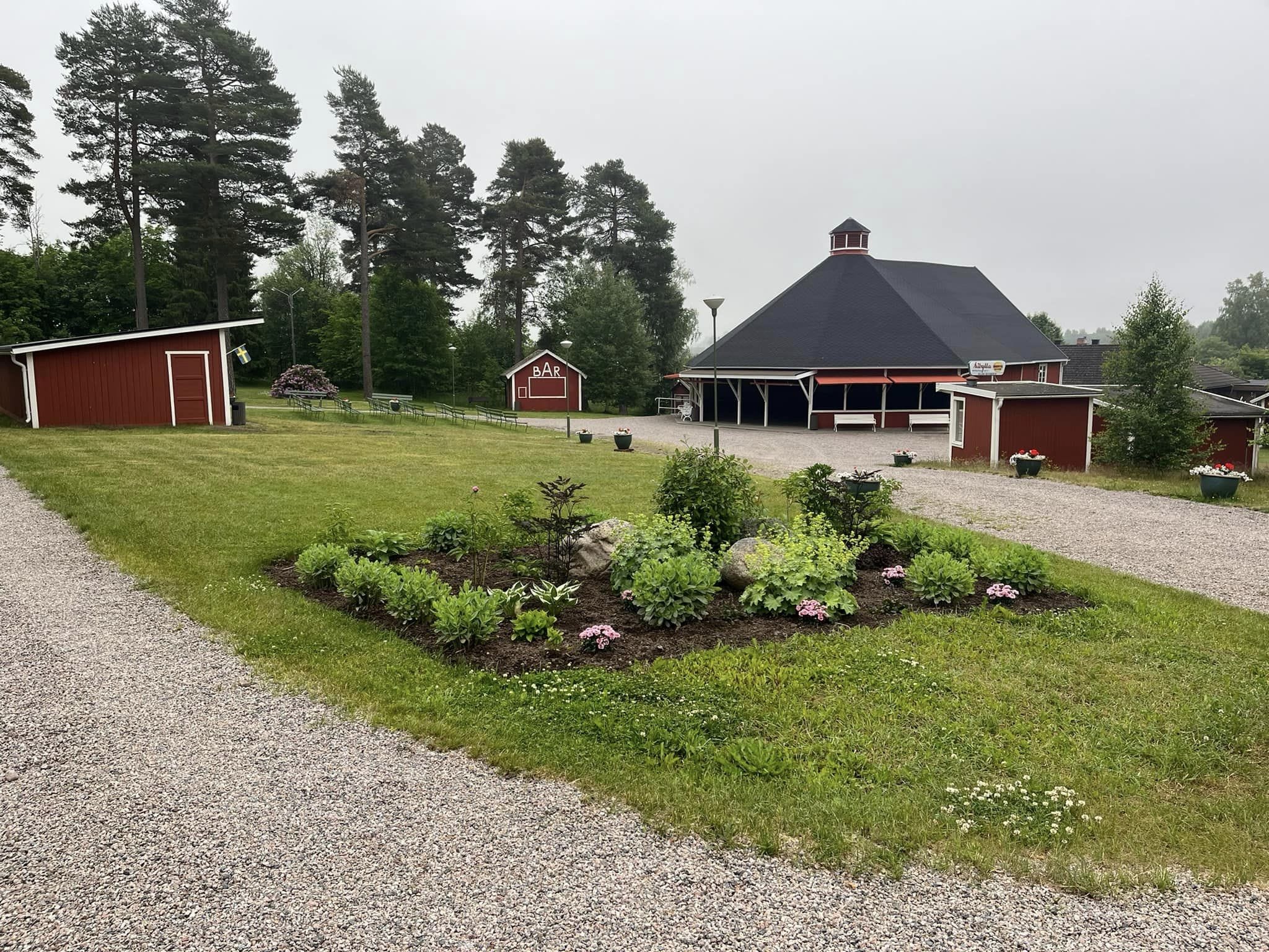 Röda byggnader kring en gräsmatta med plantering.