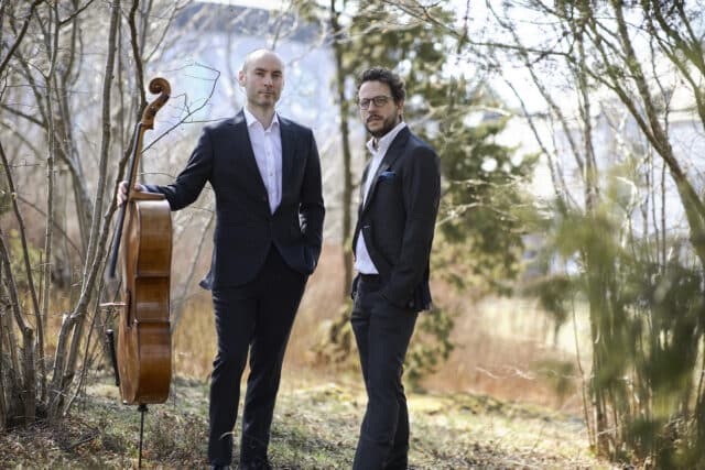 Två män i svart kostym i naturmiljö, den ena hållande en cello.