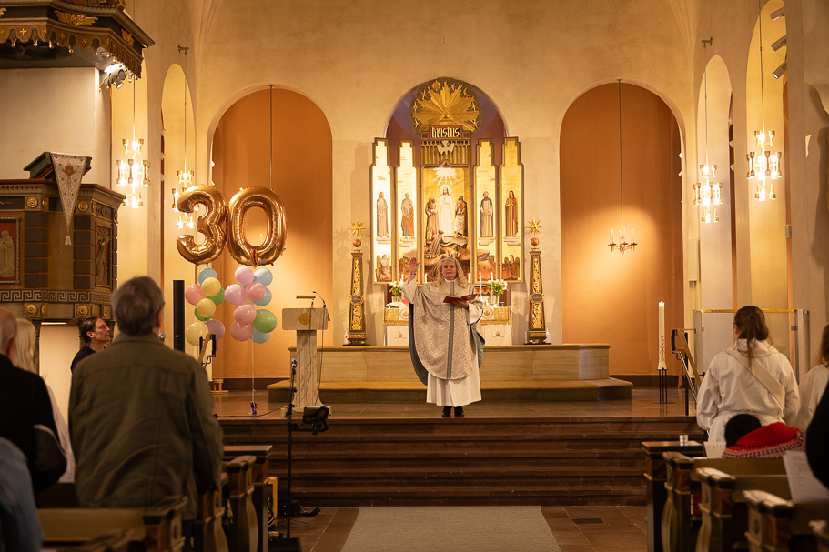 Översiktsbild av kvinnlig präst som håller gudstjänst vid altaret och där ryggarna syns på några av besökarna.