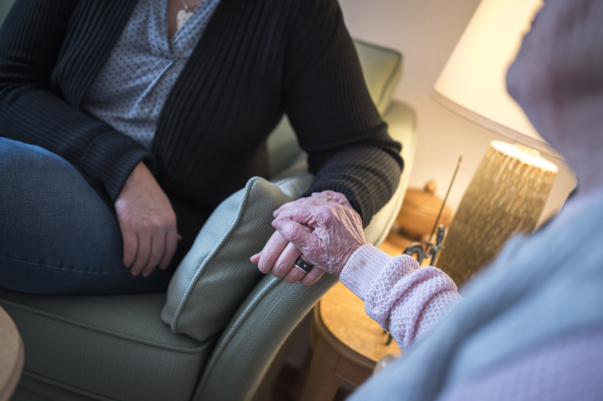 En diakon håller en äldre person i handen. De sitter båda i fåtöljer i ett vamt upplyst rum. Det är armarna och händerna som visas, inte deras ansikten.