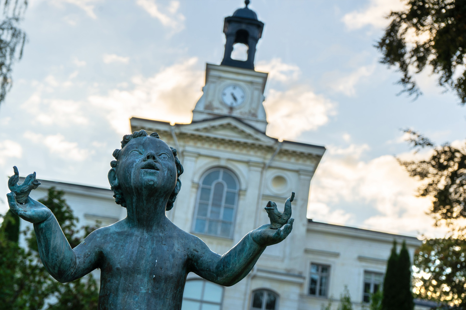En staty föreställande ett barn med två fiskar i händerna ses i förgrunden. I bakgrunden syns Gode Herdens kapell.