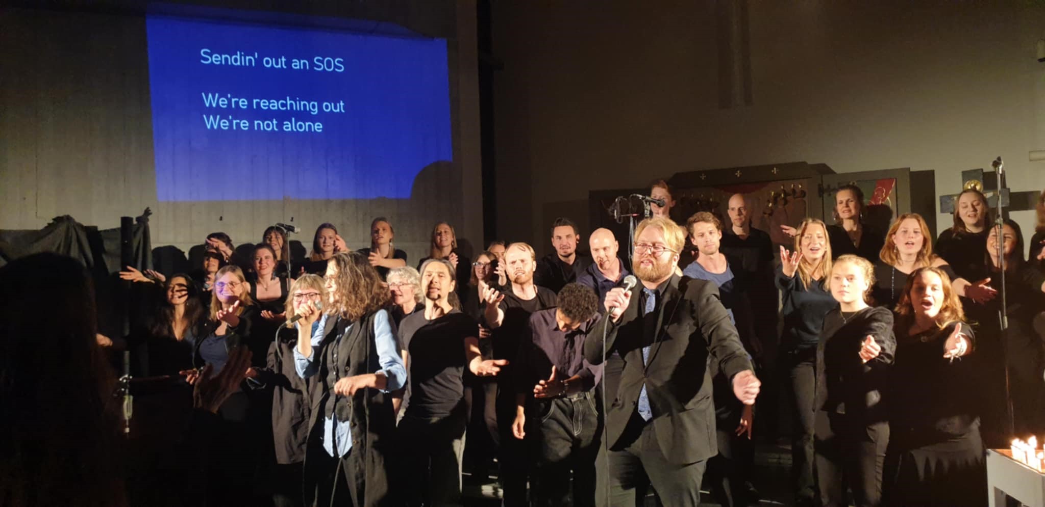 Kören EnEndaKropp syns sjungandes och dnasandes i S:t Pers kyrka med en blå textskylt projicerad på väggen snett bakom.
