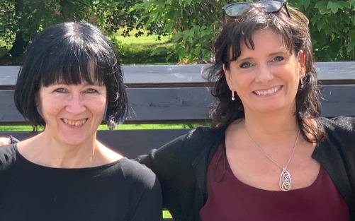 Cajsa Ekstav och Ditte Andersson sitter på en bänk i solen och ler mot kameran.