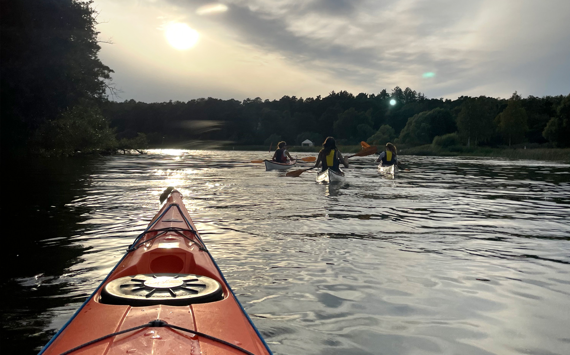 En sjö med tre kajakpaddlare. I förgrunden syns fören på fotografens kajak.