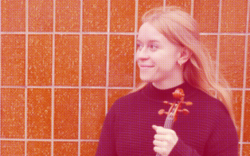 Frida Östblom står med fiol i handen mot en kakelvägg.