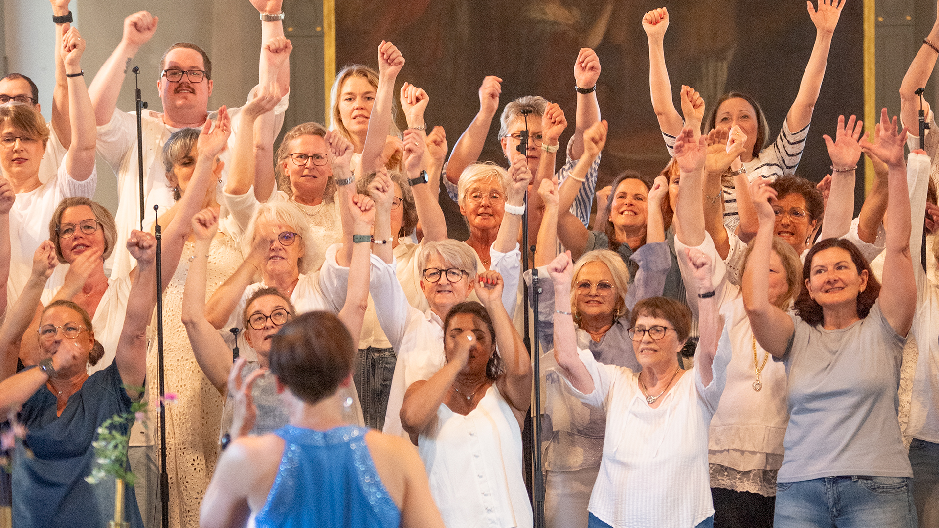 Man ser kören i ljusa kläder med uppsträckta händer. Framför står körledaren med ryggen emot, i himmelsblått.