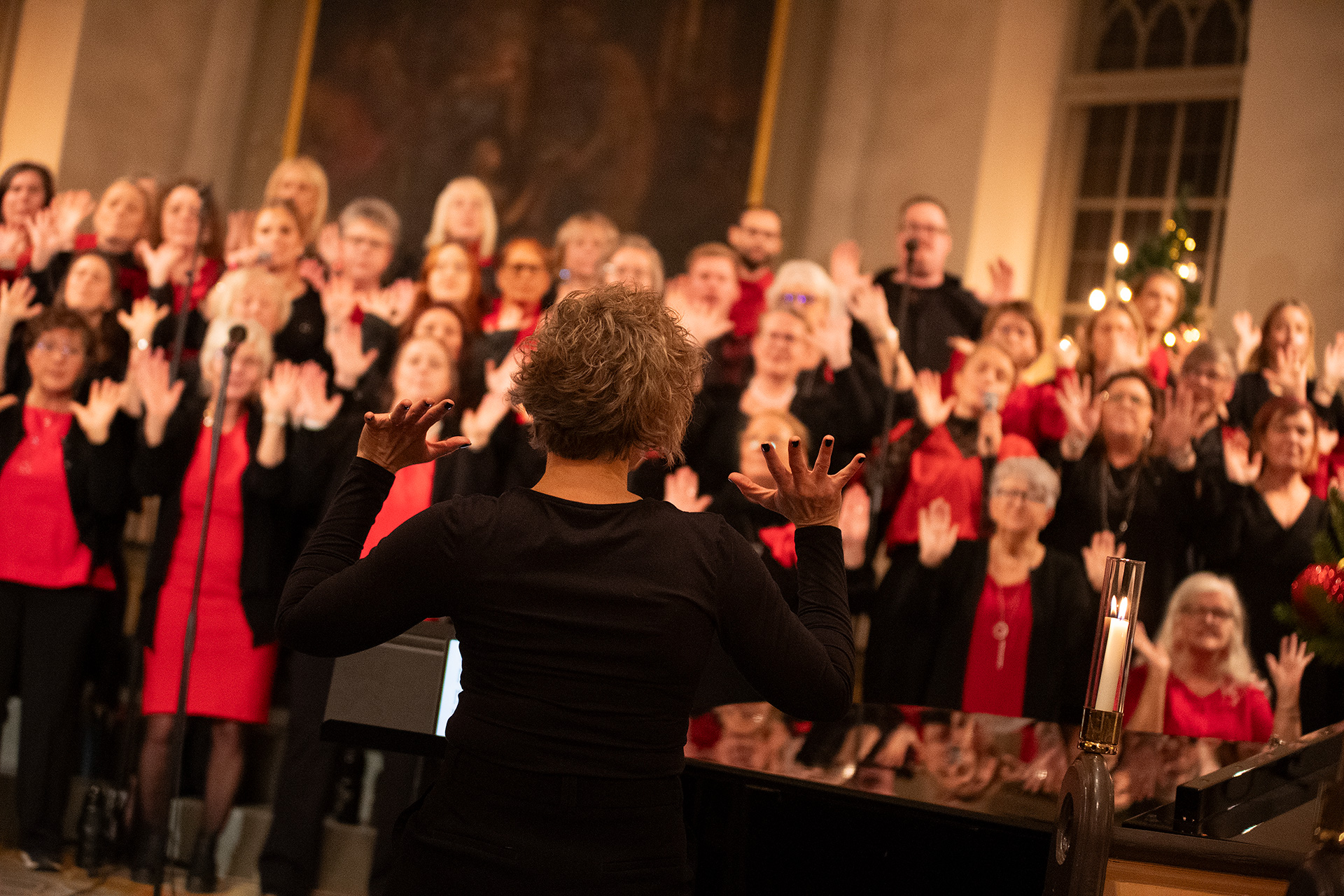 I förgrunden ser man hur körledaren dirigerar, med spretande fingrar och svarta kläder. I bakgrunden syns kören i färgerna rött och svart.