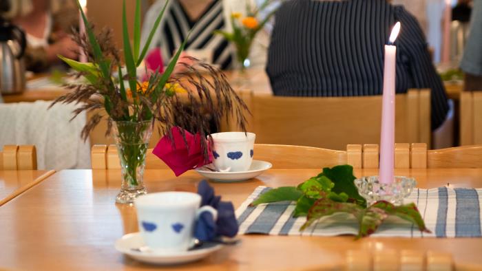 Ett uppdukat bord syns i bild. På bordet står brinnande ljusrosa ljus, vaser med gröna växter och blåvita kaffekoppar med servetter instuckna i örat. I bakgrunden syns kvinnor som samtalar och dricker kaffe.