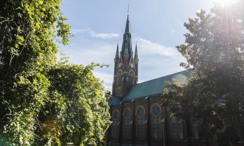 Matteus kyrka, en stor röd tegelbyggnad, sedd bakifrån. Till vänster i bild finns stora gröna träd och marken är grön med gräs.