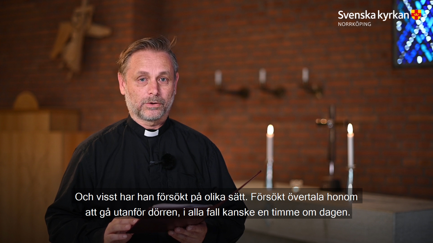En präst predikar mot en bakgrund av rött tegel och två ljus som brinner och ett kors på ett altare i bakgrunden.