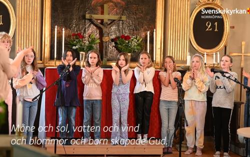 Vy över ett kor i kyrkan där en barnkör står uppställd på rad och sjunger och gör rörelser till musiken.