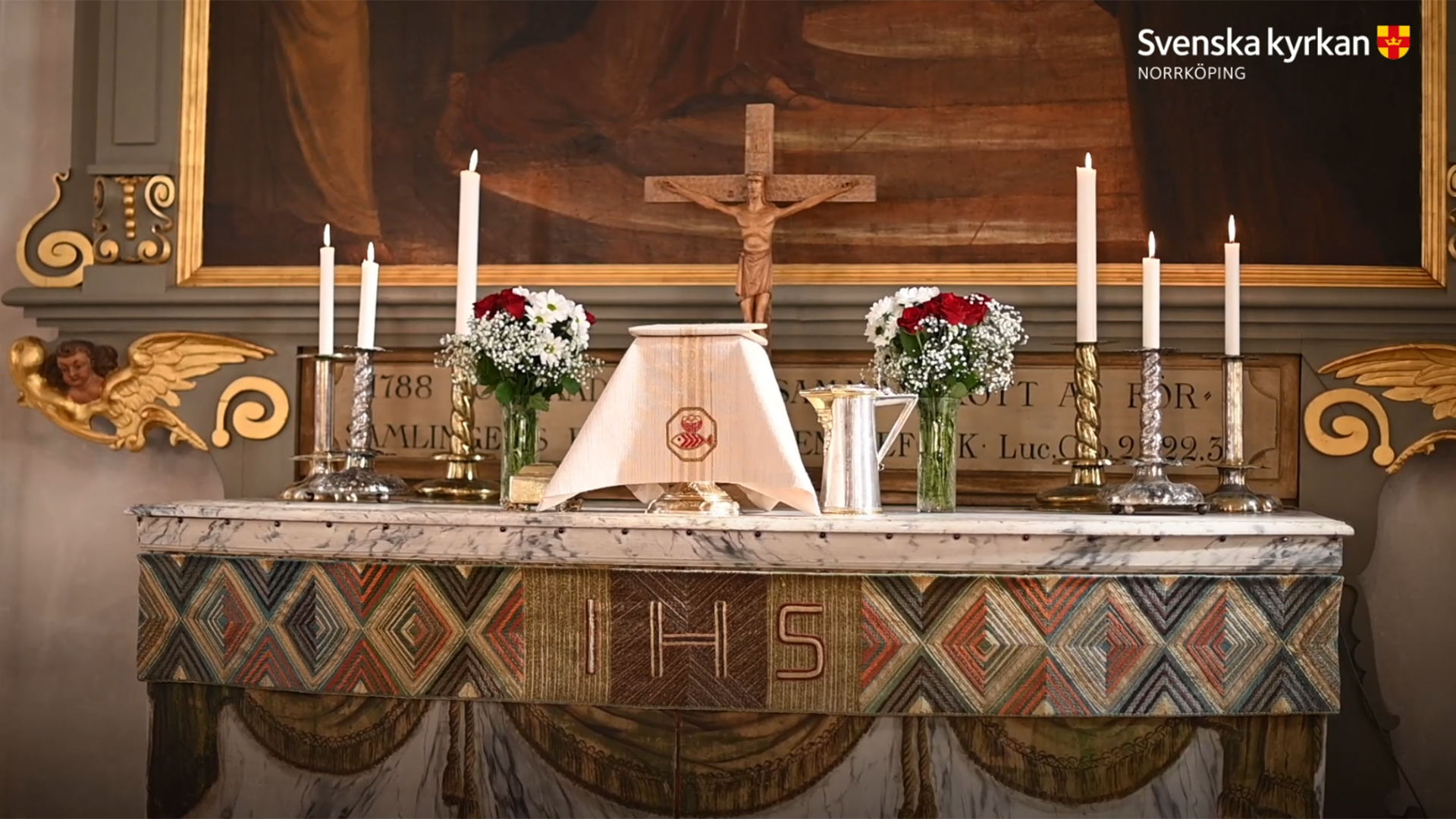 Ett altare med nattvard framdukad, sex altarljus och röda och vita blommor, som rosor och brudslöja. Kruxifixet står centrerat längst bak och altartavlan skymtar i bakgrunden.