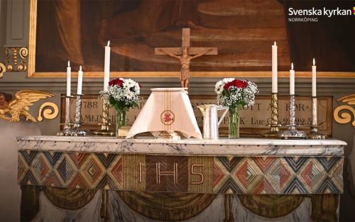 Ett altare med nattvard framdukad, sex altarljus och röda och vita blommor, som rosor och brudslöja. Kruxifixet står centrerat längst bak och altartavlan skymtar i bakgrunden.