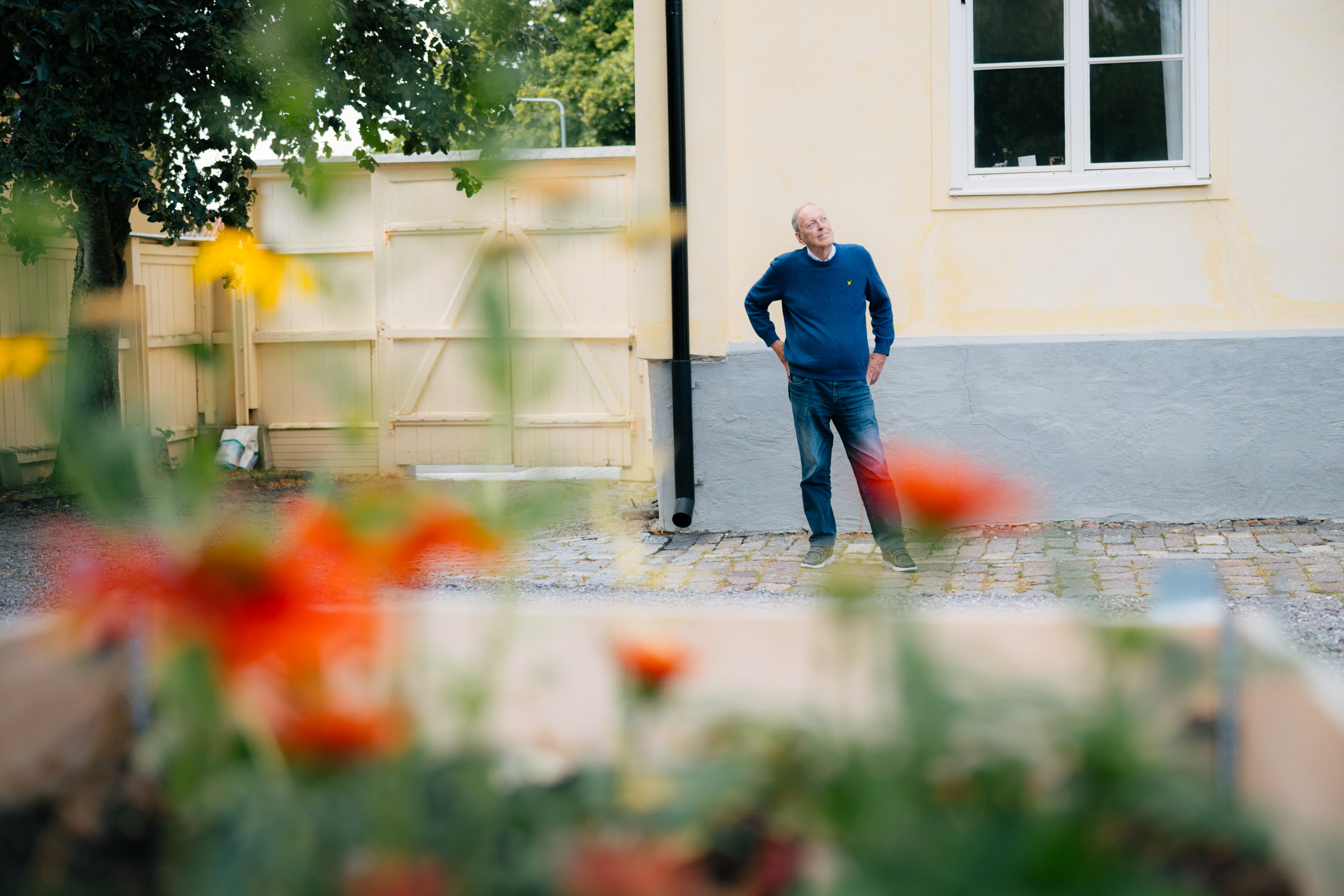 En äldre man står framför ett ljusgult putsat hus. Man ser honom på avstånd. I förgrunden syns röda vallmoblommor.