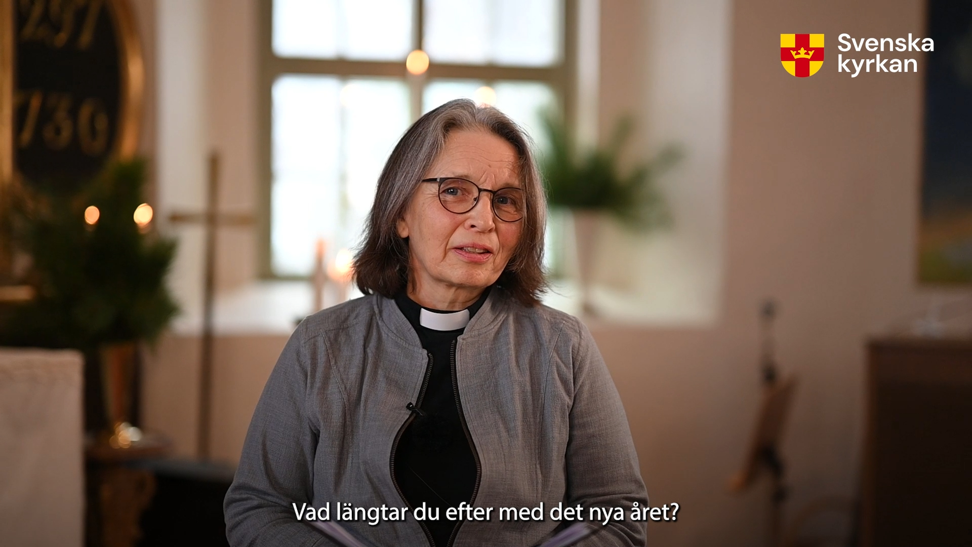 Präst Eva Strannelid står och talar med kyrkans interiör i bakgrunden. Ett fönster, ett kors, ljus i oskärpa.