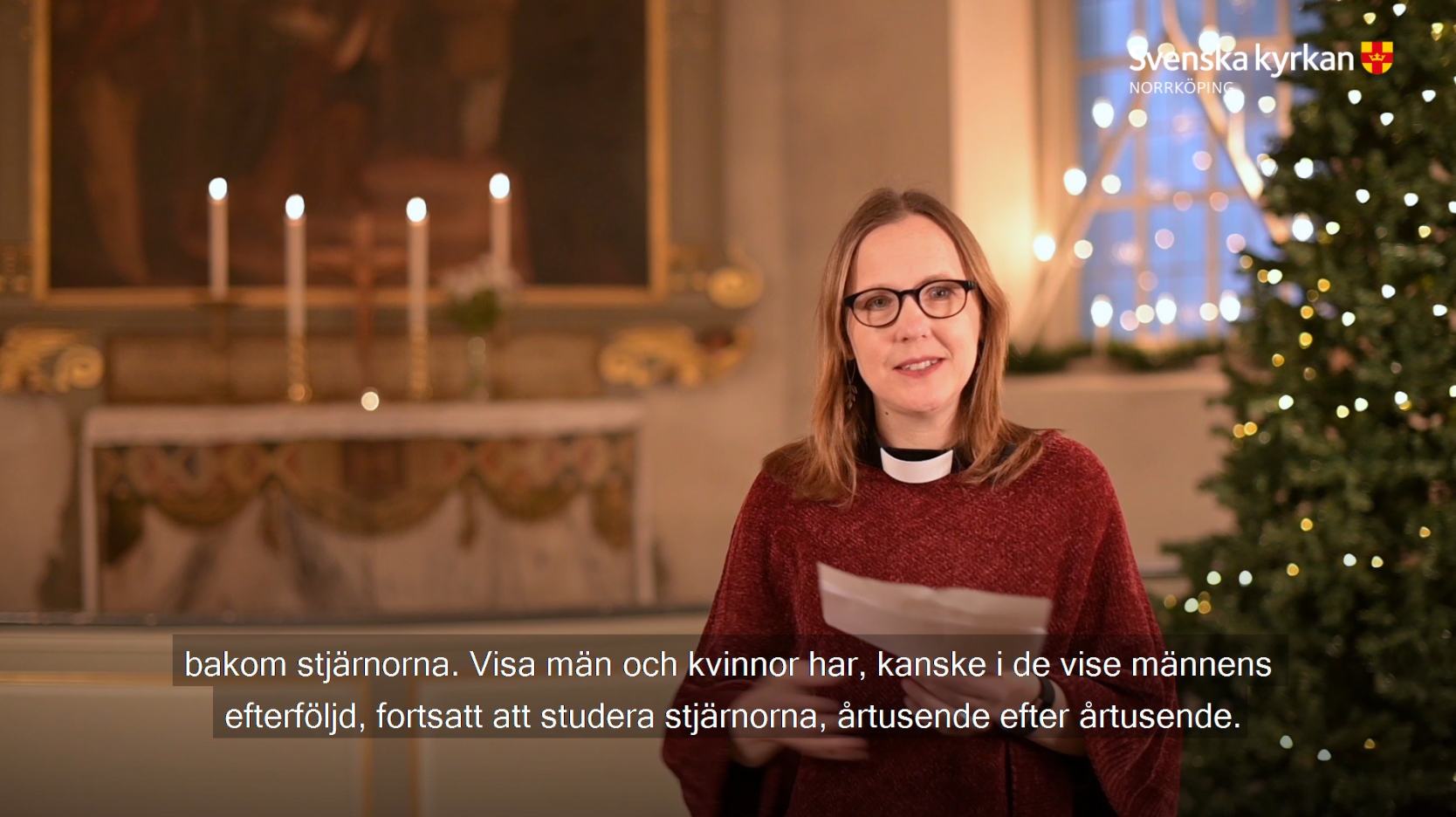 Präst Katarina står och predikar framför ett altare med tända ljus och en julgran.
