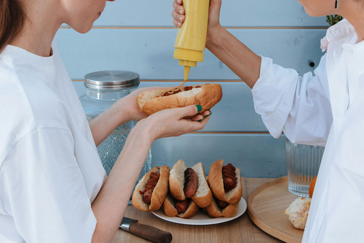 Två personer tar senap på en grillad korv med bröd. Fler korvar ligger på ett bord.