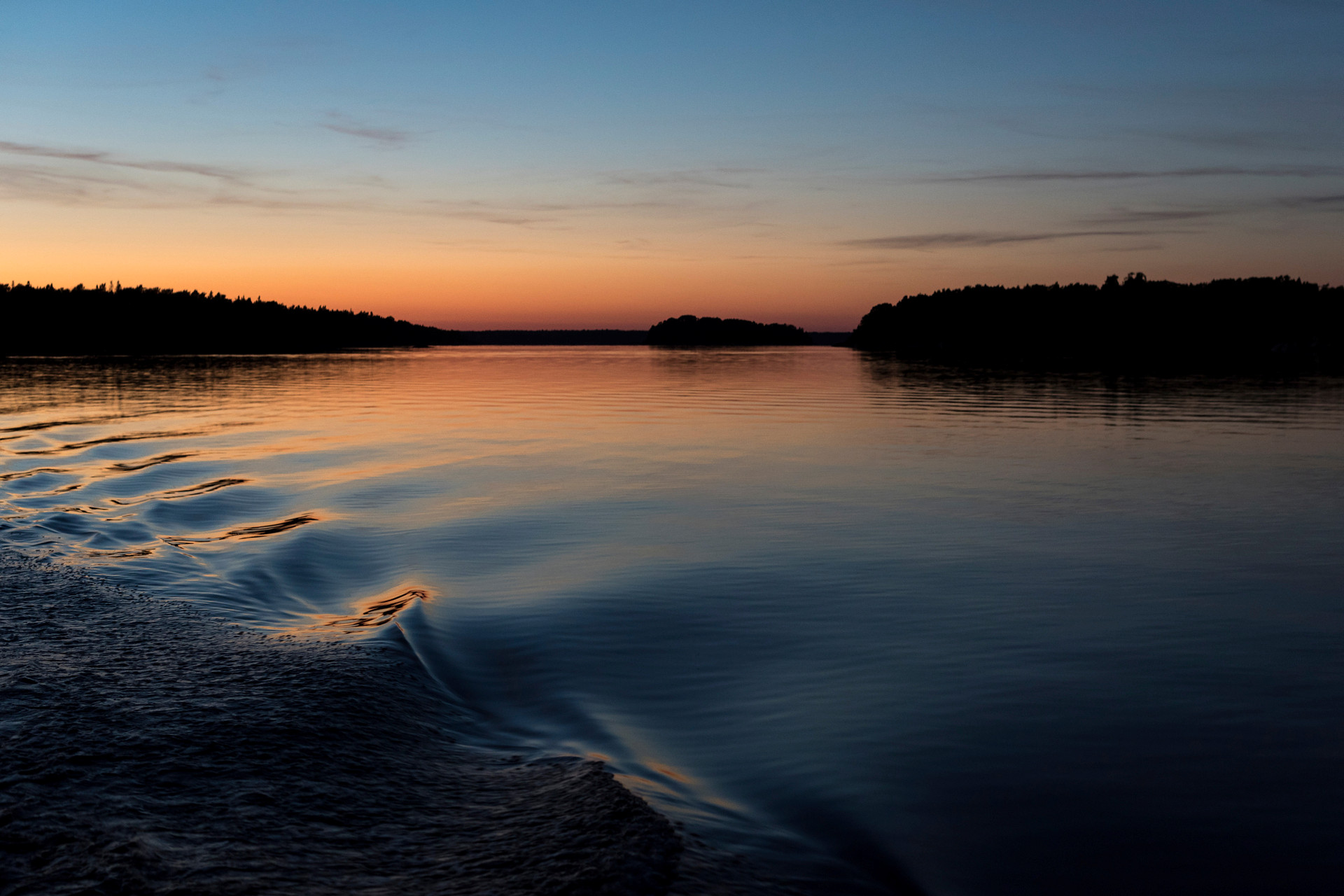 Lugna vågor på vattenytan. I bakgrunden syns skuggor av träd, solnedgång.