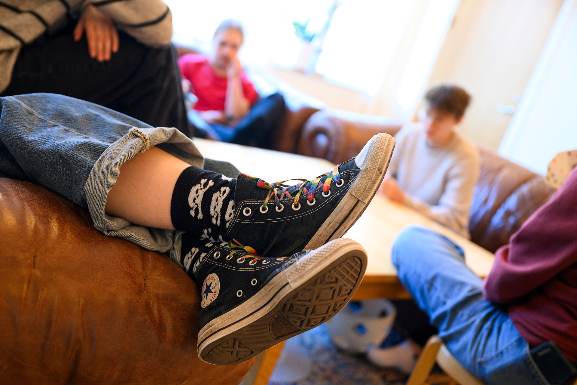 En grupp av ungdomar hänger i soffor. I förgrunden syns ett par skor med regnbågsskosnören i närbild.