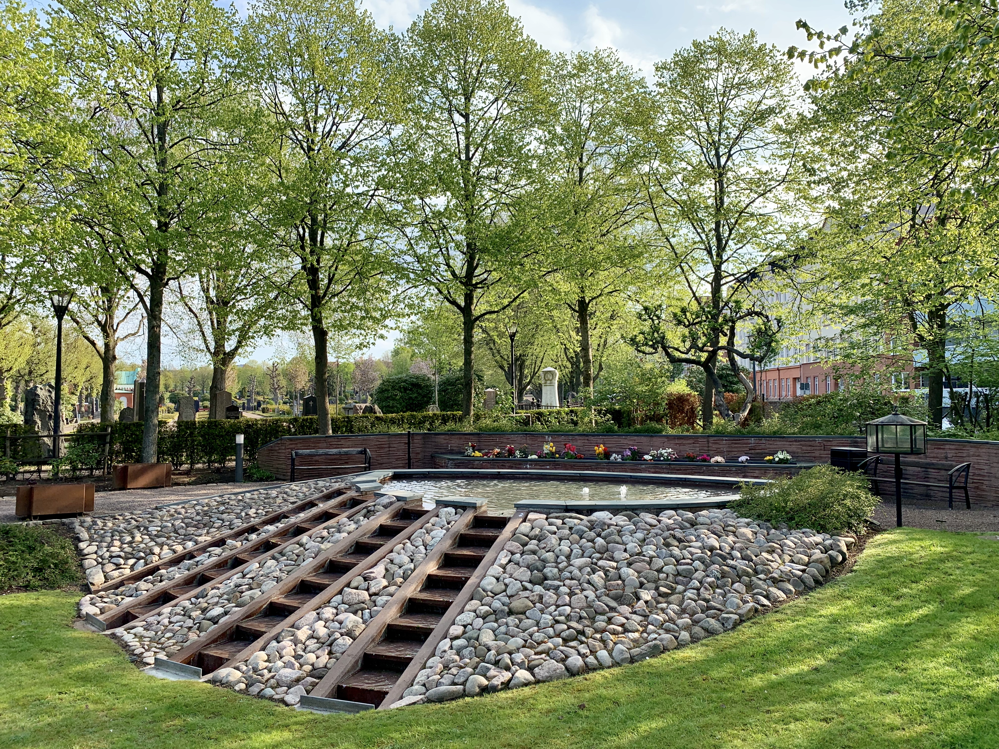 Ett stort vattenspel uppbyggt av natursten. I bakgrunden stora gröna träd samt plats för smyckning med snittblommor och värmeljus.