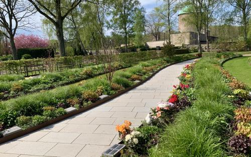 Gravrader med kalkstensplattor. Omkring gravplatser i perennplanteing. I bakgrunden Krematoriet, en brun tegelbyggnad med grönt tak. 