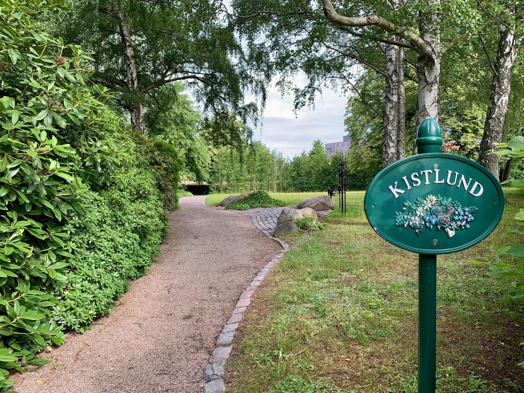 En grusgång med omkringliggande rhododendronbuskar och stora gröna träd. Intill en ängsyta står ljus- och blomsterhållare i metall.