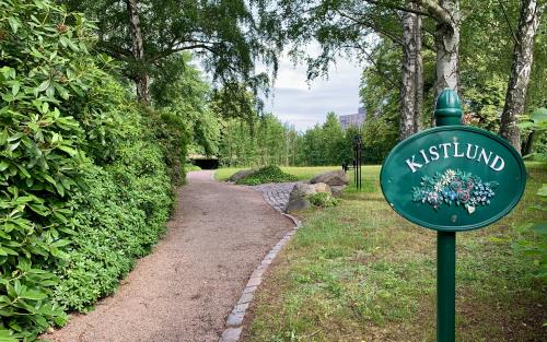 En grusgång med omkringliggande rhododendronbuskar och stora gröna träd. Intill en ängsyta står ljus- och blomsterhållare i metall.