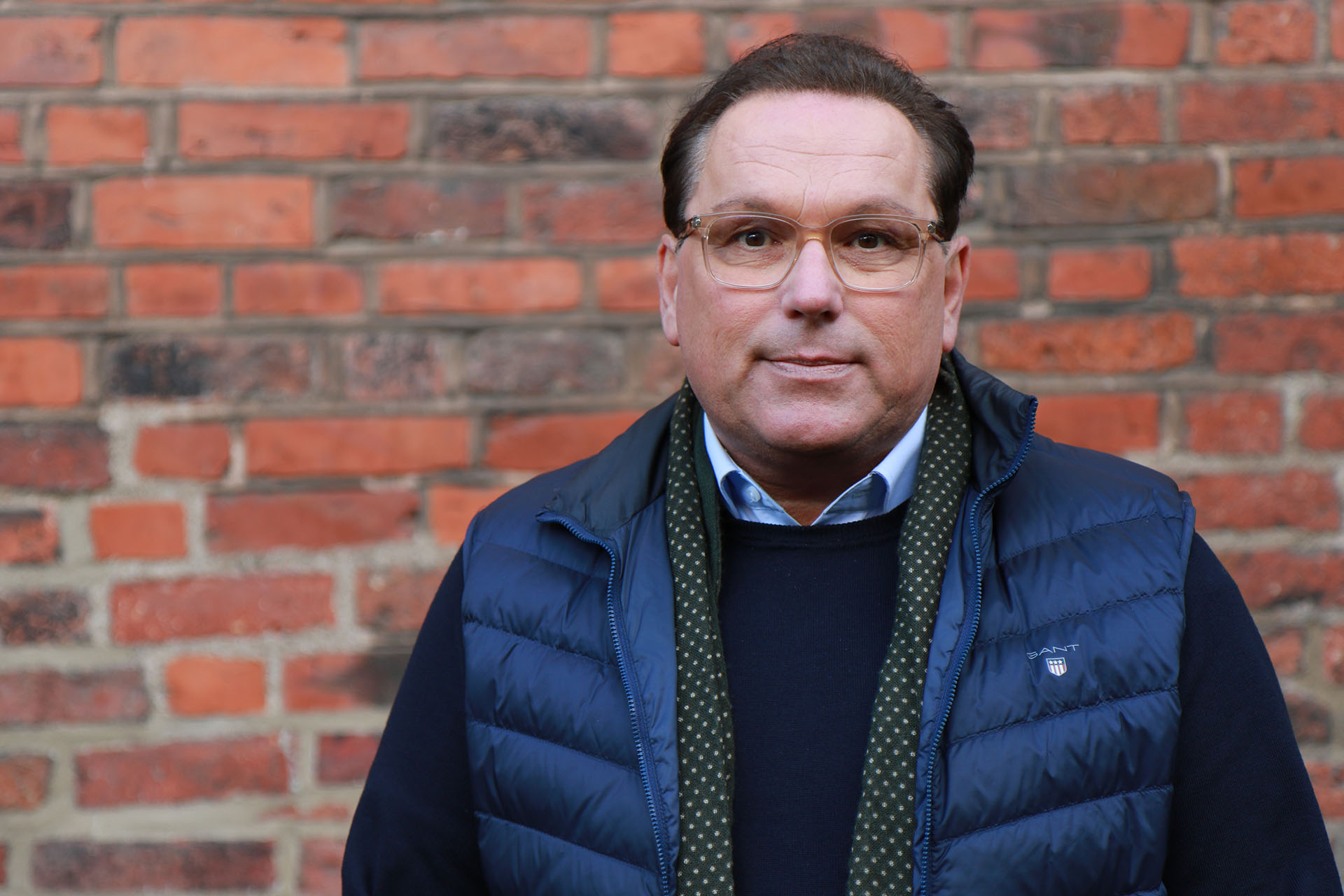 Närbild på en man i mörkt hår, klädd i en blå täckväst. I bakgrunden syns en röd tegelvägg.