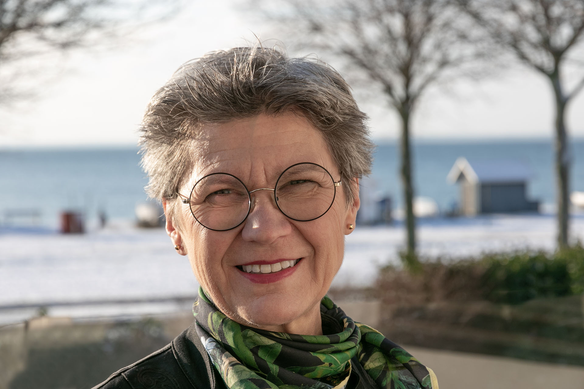 Närbild på en kvinna med kort grått hår, glasögon och en grön scarf. I bakgrunden syns en väg, havet och några träd.